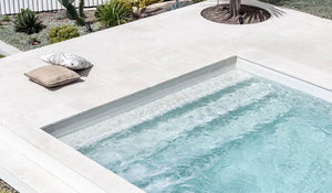 A swimming pool with blue tiles inside the pool and light beige marble tiles around the pool. It is a sunny day.